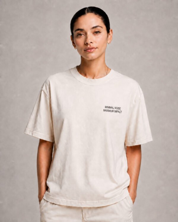 Krush Essentials 8 Model wearing a Natural Raw oversized Kat Krush T-shirt with small “Minimal Noise Maximum Impact” chest print, styled with light trousers against a clean grey studio background.
