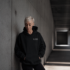 Editorial lifestyle image of female model wearing oversized black Kat Krush hoodie in minimalist concrete corridor with calm cinematic streetwear mood.