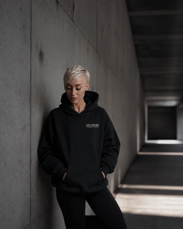 Editorial lifestyle image of female model wearing oversized black Kat Krush hoodie in minimalist concrete corridor with calm cinematic streetwear mood.
