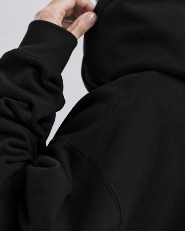 Close-up detail shot of a heavyweight black hoodie showing premium shoulder seam construction, ribbed cuff texture, and soft brushed fabric finish. Minimal studio lighting highlights the quality stitching, dense material, and oversized streetwear craftsmanship.