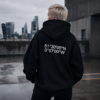 Cinematic rooftop lifestyle image featuring a distant model in a black oversized coordinates hoodie walking across wet concrete. Dark urban skyline, reflective pavement, and muted tones create a quiet confidence streetwear mood with the product subtly integrated into the scene.