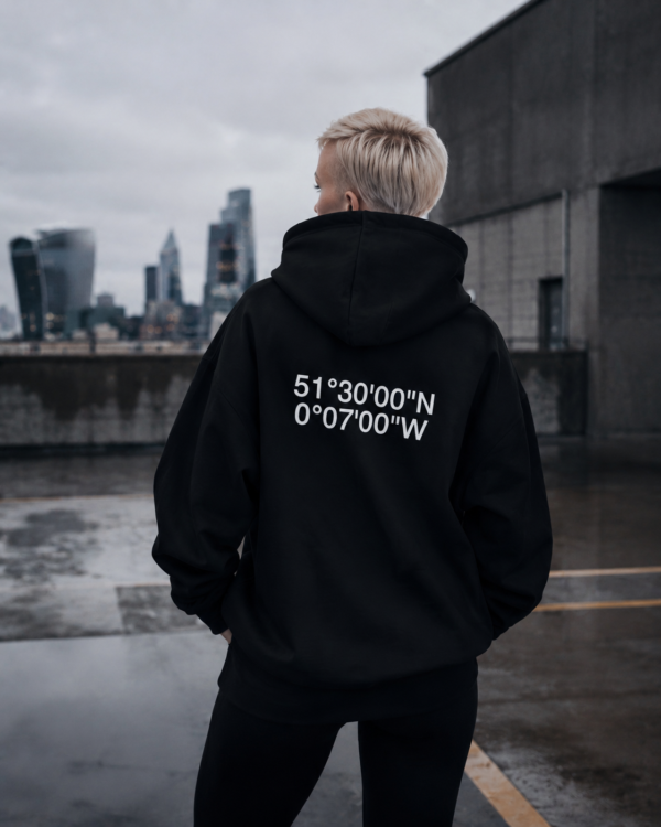 Cinematic rooftop lifestyle image featuring a distant model in a black oversized coordinates hoodie walking across wet concrete. Dark urban skyline, reflective pavement, and muted tones create a quiet confidence streetwear mood with the product subtly integrated into the scene.
