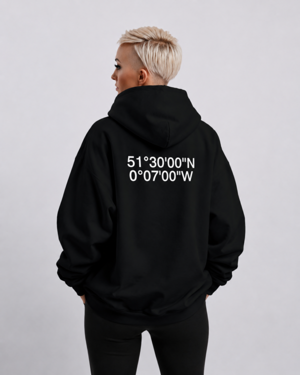 Oversized black hoodie with white London coordinates back print, shown in a relaxed waist-up pose. Model turned slightly away from camera to highlight the oversized drape, dropped shoulders, and heavyweight silhouette against a clean neutral studio background.
