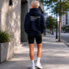 Minimal black oversized hoodie featuring “MINIMAL NOISE MAXIMUM IMPACT” back print, photographed in a calm modern city street setting with concrete architecture, soft evening light, and relaxed editorial streetwear styling.