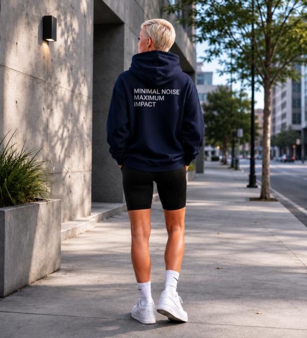 Minimal black oversized hoodie featuring “MINIMAL NOISE MAXIMUM IMPACT” back print, photographed in a calm modern city street setting with concrete architecture, soft evening light, and relaxed editorial streetwear styling.