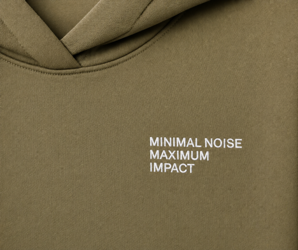 Detailed close-up of khaki hoodie chest print featuring minimalist white “MINIMAL NOISE MAXIMUM IMPACT” typography on heavyweight textured fabric with clean modern streetwear styling.