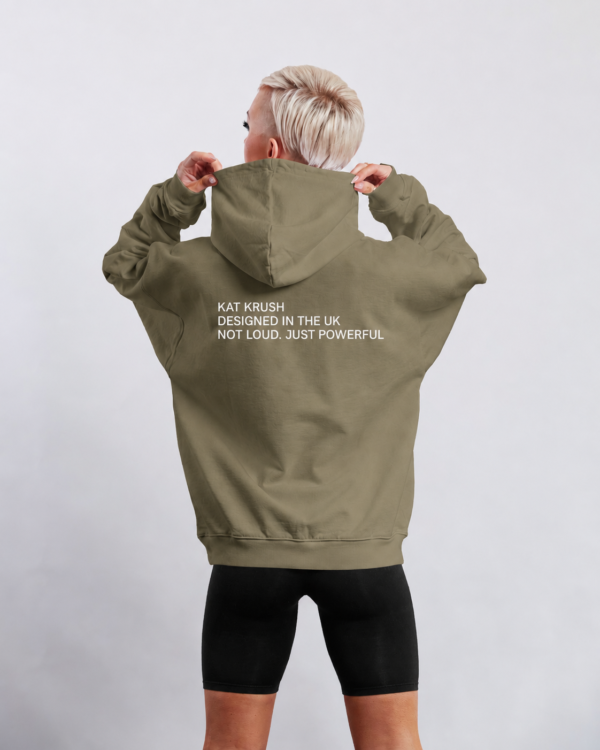 kk-hoodie-not-loud-just-powerful-back-print-khaki Back view of oversized khaki heavyweight hoodie featuring bold white “KAT KRUSH — DESIGNED IN THE UK — NOT LOUD. JUST POWERFUL” typography print, styled in a clean studio setting with relaxed minimalist streetwear aesthetic.