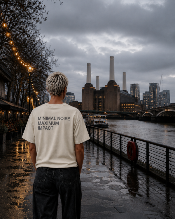 kk-tee-minimal-noise-max-impact-back-print-natural-raw-battersea Model standing on a riverside walkway near Battersea Power Station wearing a natural raw oversized T-shirt with “MINIMAL NOISE MAXIMUM IMPACT” back print. Wet pavement, city lights and overcast London skies create a calm urban editorial atmosphere.