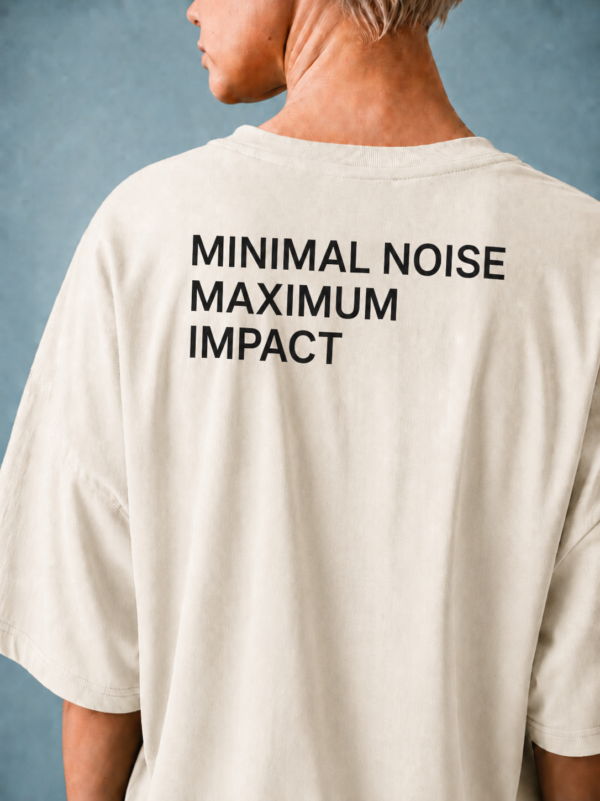 kk-tee-minimal-noise-max-impact-back-print-natural-raw1 Rear view of a model wearing a natural raw oversized T-shirt featuring the statement “MINIMAL NOISE MAXIMUM IMPACT” printed in large white lettering across the back. Soft studio lighting emphasizes the premium heavyweight cotton and relaxed silhouette.