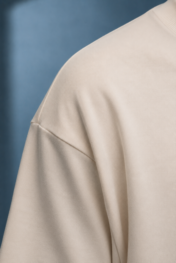 kk-tee-minimal-noise-max-impact-closeup-fabric-natural-raw Close-up of the shoulder, sleeve and ribbed collar of a natural raw oversized T-shirt, highlighting the smooth heavyweight cotton fabric, clean stitching and relaxed dropped-shoulder construction.