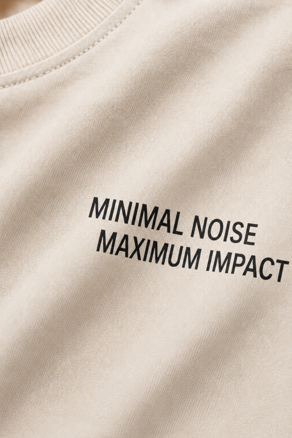 kk-tee-minimal-noise-max-impact-front-print-natural-raw Detailed close-up of the chest area of a natural raw oversized T-shirt featuring the minimalist slogan “MINIMAL NOISE MAXIMUM IMPACT” printed in black. The image showcases fabric texture, print quality and premium construction.