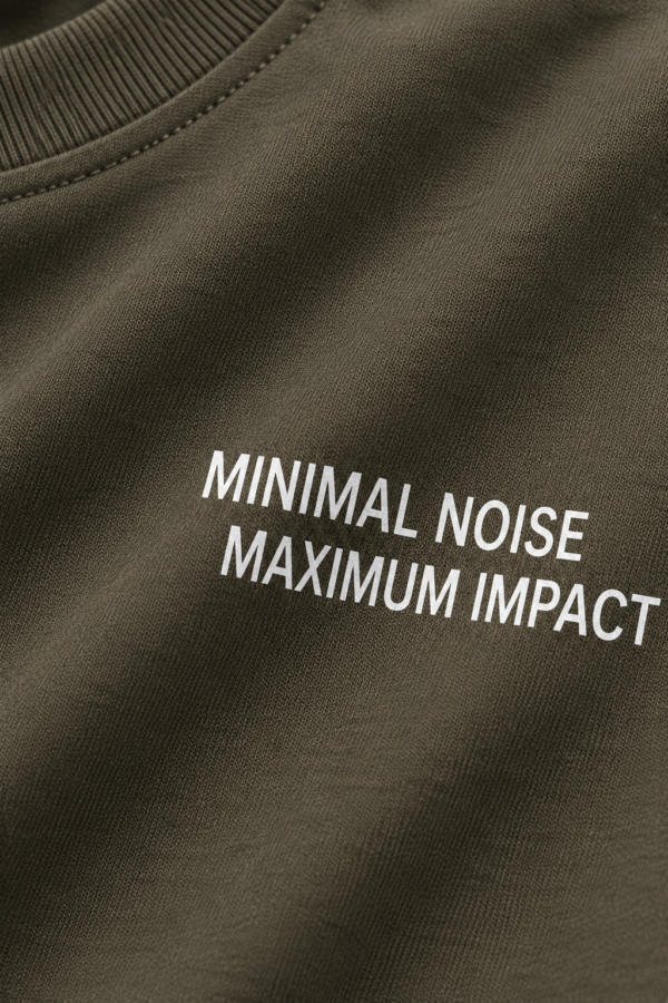 Close-up fabric detail of khaki heavyweight oversized t-shirt with “MINIMAL NOISE MAXIMUM IMPACT” typography print and visible cotton texture folds