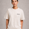 kk-tee-minimal-noise-max-impact-studio-front-natural-raw-v2 Model wearing a Natural Raw oversized Kat Krush T-shirt with small “Minimal Noise Maximum Impact” chest print, styled with light trousers against a clean grey studio background.