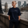 Woman wearing an oversized French navy Kat Krush t-shirt with “NOT LOUD JUST POWERFUL” chest print, standing by the River Thames near Tower Bridge in a moody London city scene with hands in pockets and an editorial streetwear aesthetic.