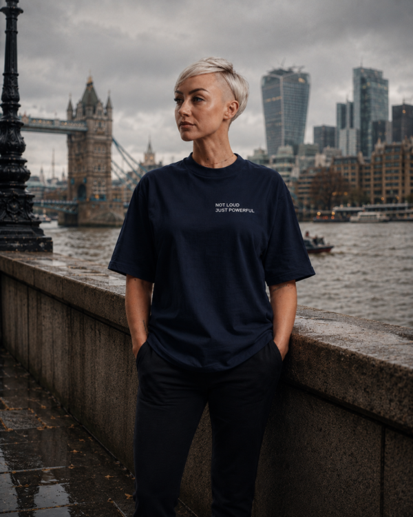 Woman wearing an oversized French navy Kat Krush t-shirt with “NOT LOUD JUST POWERFUL” chest print, standing by the River Thames near Tower Bridge in a moody London city scene with hands in pockets and an editorial streetwear aesthetic.