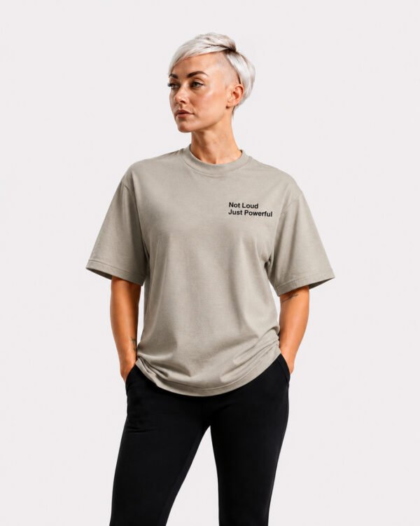 Model wearing a stone-coloured oversized Kat Krush T-shirt with “Not Loud Just Powerful” chest print, standing casually with hands in pockets against a light grey studio background.