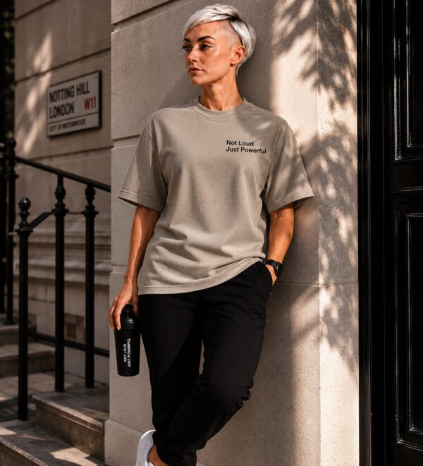 Model wearing a stone oversized Kat Krush T-shirt with “Not Loud Just Powerful” chest print, leaning against a London townhouse exterior in a quiet urban setting.