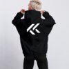 Black oversized hoodie with large white Kat Krush KK logo printed on the back. Model stands facing away while holding the hood against a clean light grey studio background.