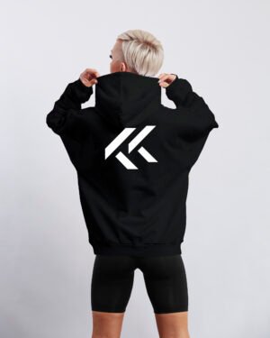 Black oversized hoodie with large white Kat Krush KK logo printed on the back. Model stands facing away while holding the hood against a clean light grey studio background.