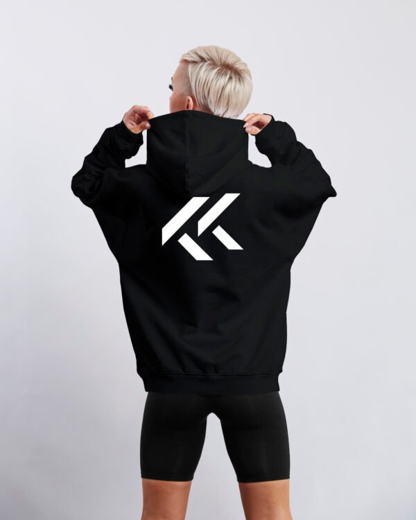 Black oversized hoodie with large white Kat Krush KK logo printed on the back. Model stands facing away while holding the hood against a clean light grey studio background.