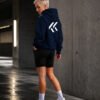 Model wearing a black Kat Krush oversized hoodie with large white KK back print in a modern concrete urban setting, showcasing minimalist streetwear style.