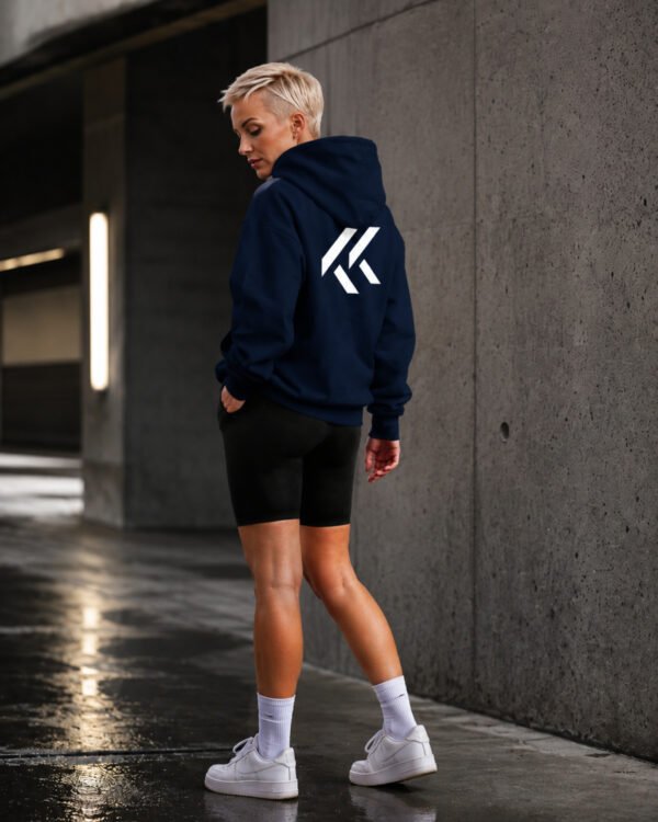 Model wearing a black Kat Krush oversized hoodie with large white KK back print in a modern concrete urban setting, showcasing minimalist streetwear style.