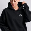 Female model wearing a black oversized Kat Krush hoodie with small white KK chest logo. Studio portrait showcasing the relaxed fit, kangaroo pocket, and minimalist branding.
