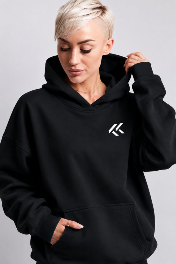 Female model wearing a black oversized Kat Krush hoodie with small white KK chest logo. Studio portrait showcasing the relaxed fit, kangaroo pocket, and minimalist branding.