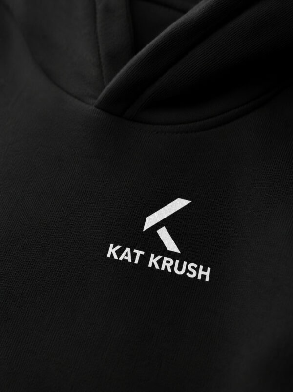 Close-up of black Kat Krush oversized hoodie showing white chest logo print and heavyweight cotton fabric texture. Premium minimal streetwear detail.