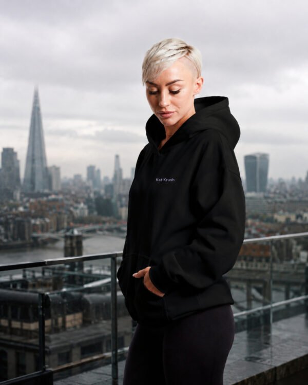 Black oversized Kat Krush hoodie with lilac chest logo styled on a London rooftop overlooking the city skyline. Minimal streetwear aesthetic with relaxed oversized fit.