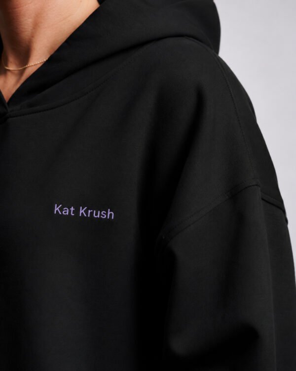 Detailed close-up of lilac Kat Krush chest logo on black oversized hoodie. Premium heavyweight cotton fabric with clean minimal branding and dropped shoulder construction.