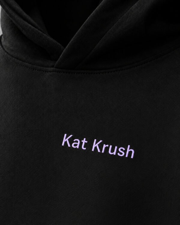 Close-up of black oversized hoodie featuring subtle lilac Kat Krush chest logo. Heavyweight cotton fabric with premium minimal streetwear styling.