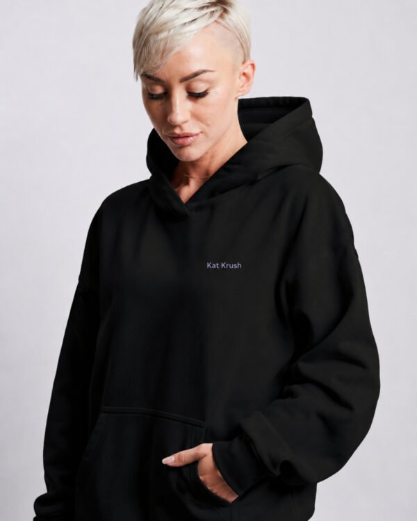 Black oversized Kat Krush hoodie with subtle lilac chest logo. Relaxed heavyweight fit with kangaroo pocket and contemporary minimal streetwear silhouette.