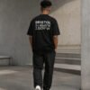 kk-tee-bristol-black-back-concrete-city-view Model wearing a black Bristol coordinates T-shirt walking through a minimalist concrete architectural setting, showcasing the oversized fit and back print design.