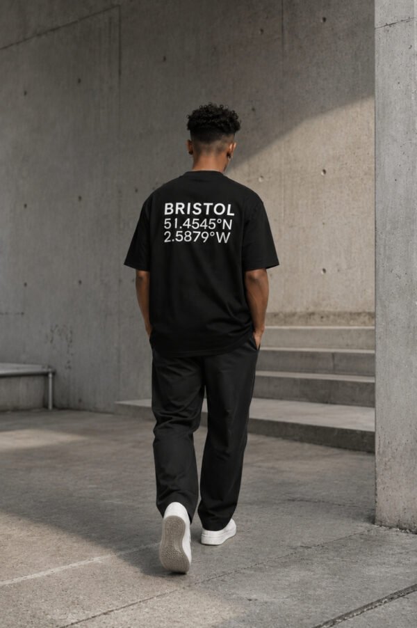 kk-tee-bristol-black-back-concrete-city-view Model wearing a black Bristol coordinates T-shirt walking through a minimalist concrete architectural setting, showcasing the oversized fit and back print design.