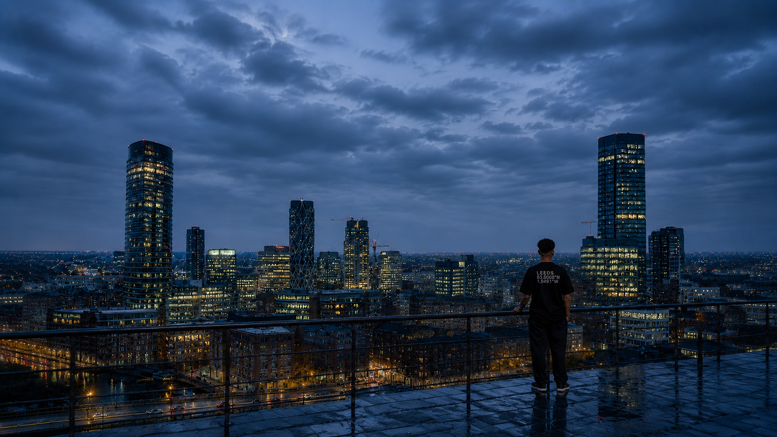 Male model wearing a black Kat Krush Leeds Coordinates oversized T-shirt overlooks the Leeds skyline from a rooftop at soft silver twilight, framed by modern glass skyscrapers, subtle blue reflections and calm editorial streetwear styling.