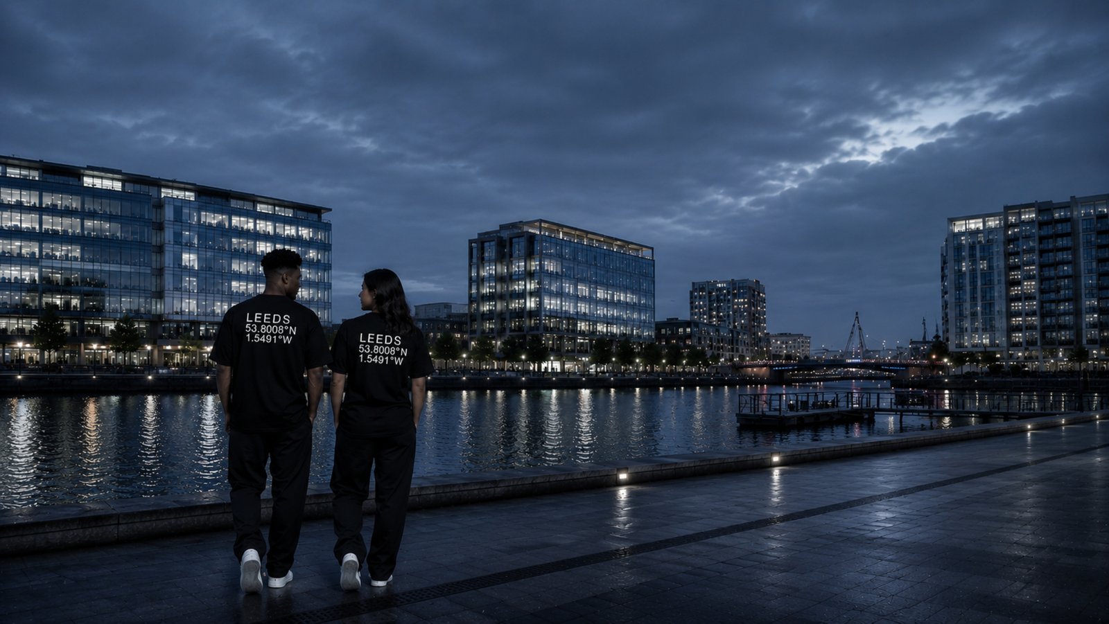 Male and female models wearing black Kat Krush Leeds Coordinates oversized T-shirts walk beside Leeds Dock at soft silver twilight, surrounded by contemporary glass architecture and calm waterfront reflections in a premium editorial streetwear scene.