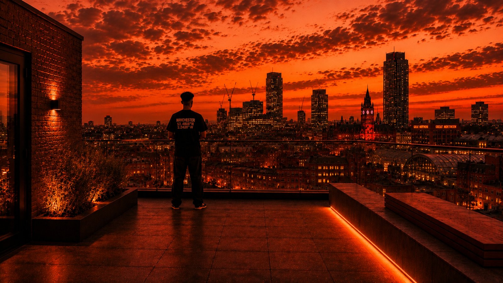 Person wearing a black Manchester Coordinates T-shirt by Kat Krush on a rooftop overlooking the Manchester skyline at sunset.
