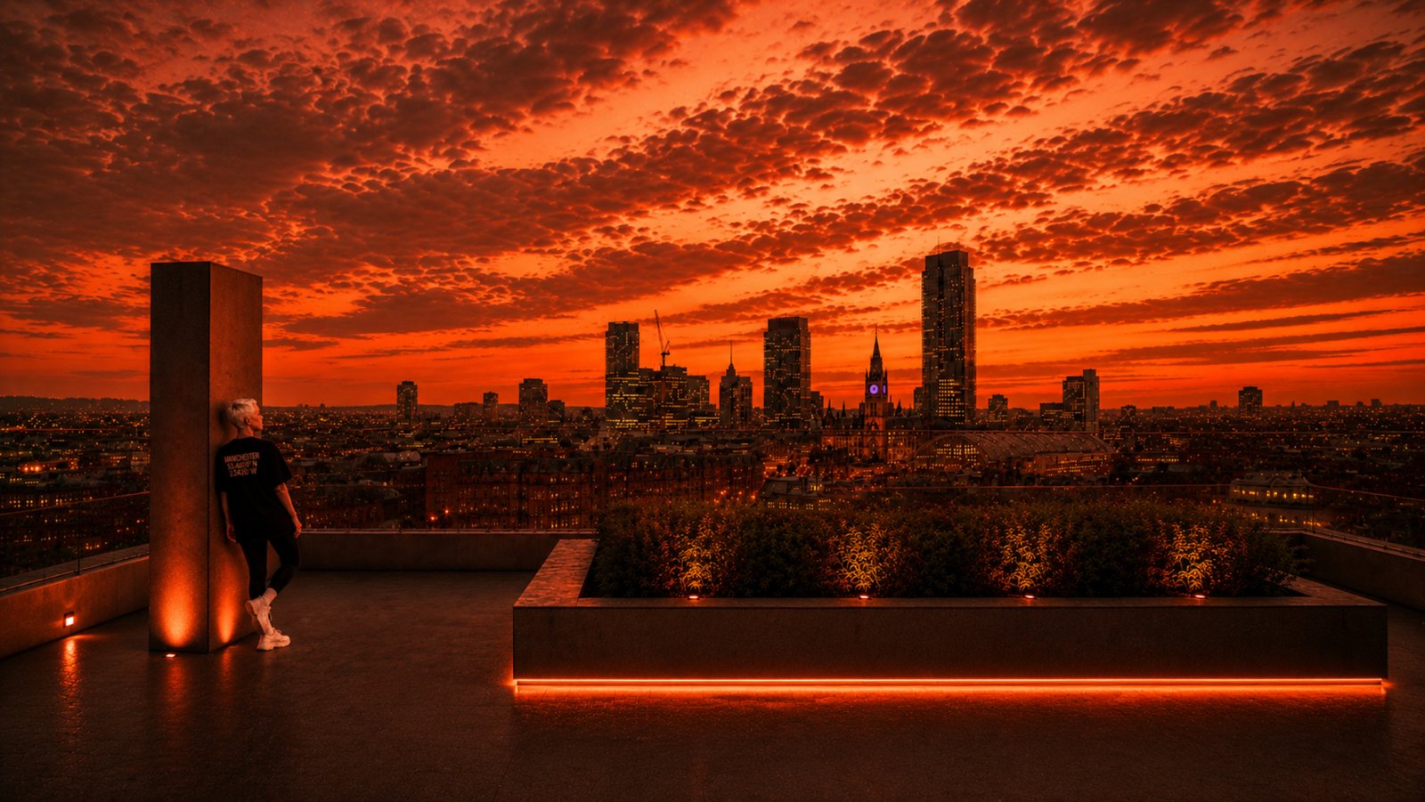 A person with short light hair leans against a rooftop pillar while looking across the Manchester skyline at sunset. They are wearing a black T-shirt with “MANCHESTER” and city coordinates printed on the back. Warm lighting along the terrace contrasts with the glowing city buildings and colourful evening sky.