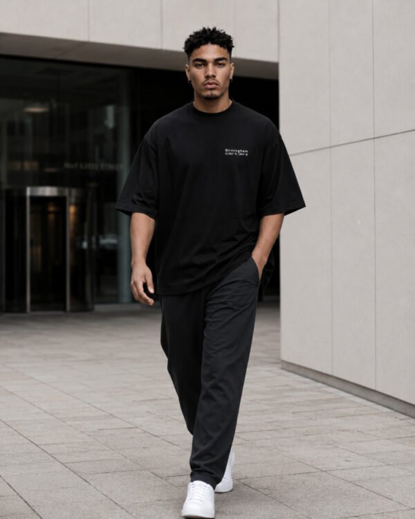 Male model walking through contemporary Birmingham-inspired architecture wearing a black Birmingham Signature oversized heavyweight T-shirt with minimalist white chest print.