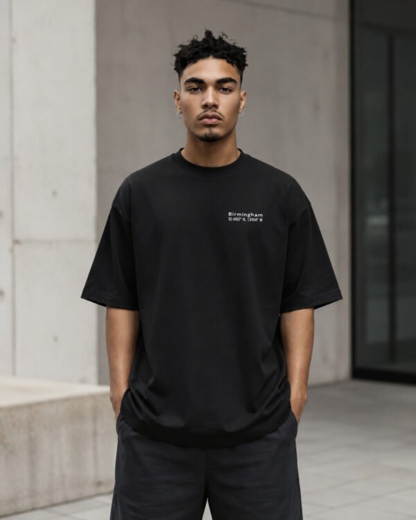 Male model wearing a black Birmingham Signature oversized heavyweight T-shirt with white chest coordinates print against contemporary Birmingham-inspired architecture.