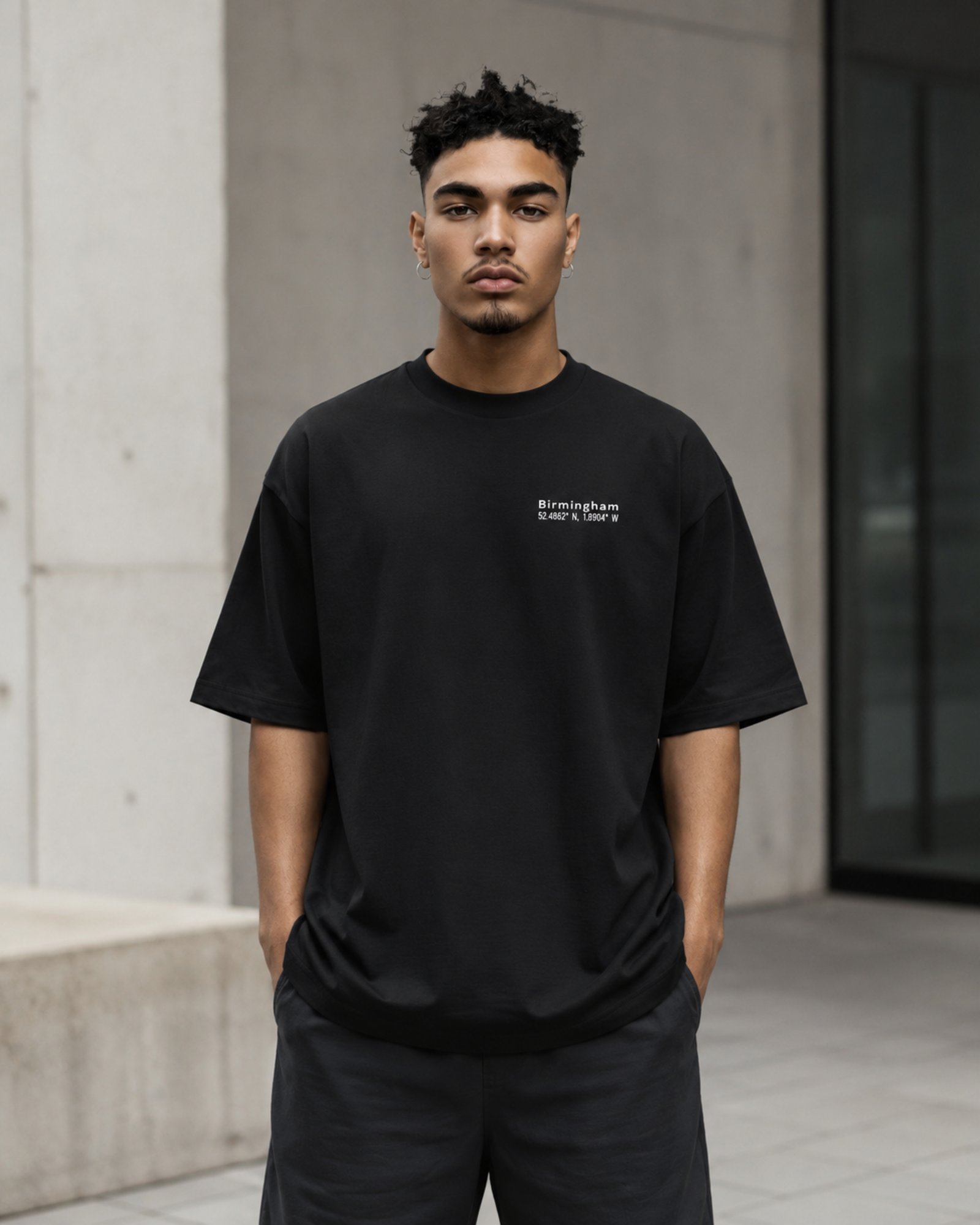 Side profile of a male model wearing a black Birmingham Signature oversized heavyweight T-shirt with relaxed fit and subtle white chest coordinates design.