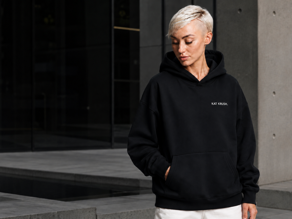 Female model wearing an oversized black Kat Krush hoodie with subtle white chest branding, standing against modern concrete and dark glass architecture.