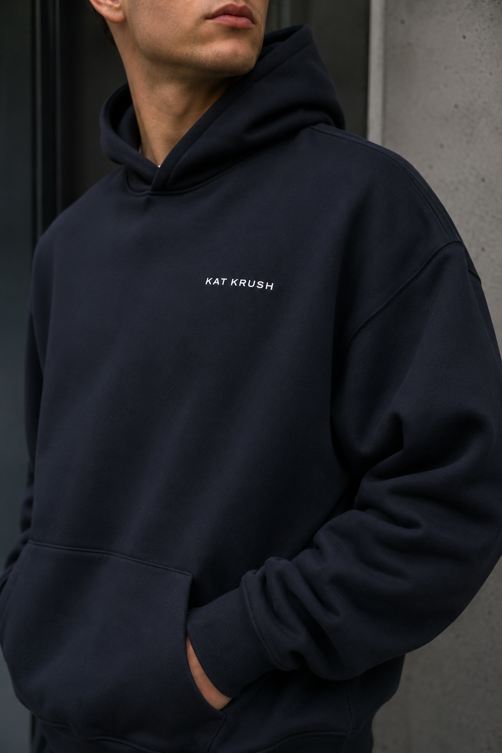 Model wearing a black oversized Kat Krush hoodie and cap beside modern concrete and glass architecture.
