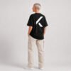 Full-length rear view of a female model wearing a black oversized Kat Krush T-shirt with a large white signature K back print and off-white cargo trousers in a minimalist studio.