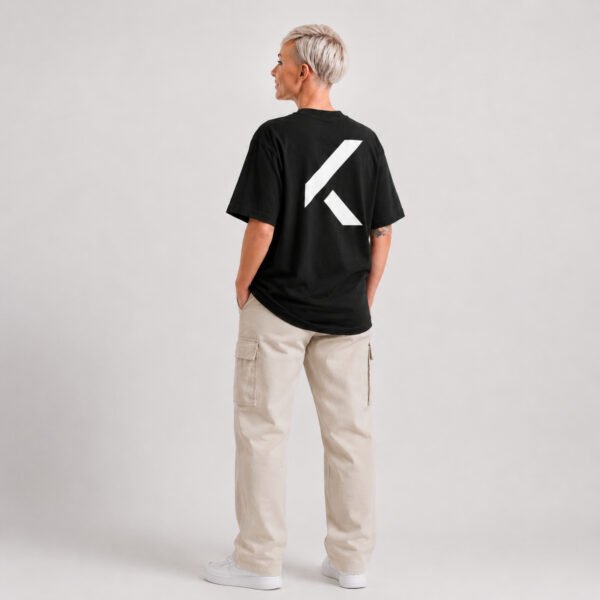 Full-length rear view of a female model wearing a black oversized Kat Krush T-shirt with a large white signature K back print and off-white cargo trousers in a minimalist studio.