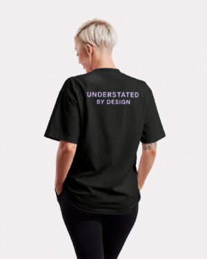 Rear view of a black Kat Krush oversized T-shirt featuring the Understated By Design statement print across the upper back in a minimalist studio setting.