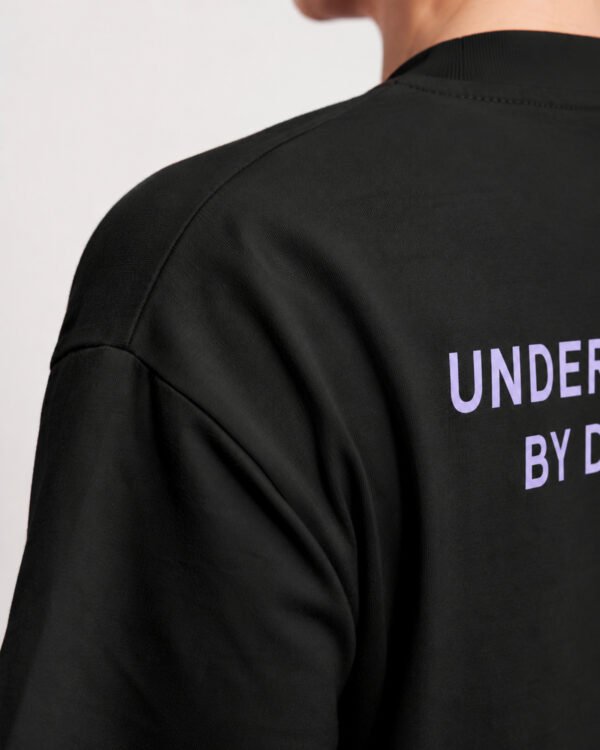 Close-up of the heavyweight cotton fabric and shoulder seam on a black Kat Krush oversized T-shirt, highlighting the Understated By Design back print.