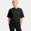 Model wearing a black Kat Krush oversized heavyweight T-shirt with minimalist chest logo, styled with black trousers in a relaxed studio portrait.