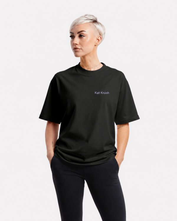 Model wearing a black Kat Krush oversized heavyweight T-shirt with minimalist chest logo, styled with black trousers in a relaxed studio portrait.